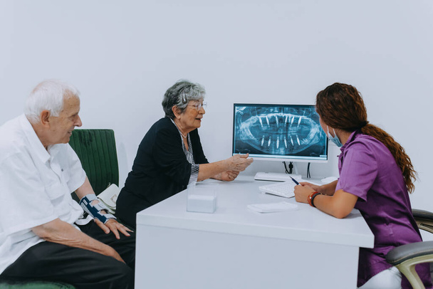 Un couple de personnes âgées discute sérieusement avec son dentiste des options de prothèse moderne dans un cabinet dentaire contemporain.. - Photo, image