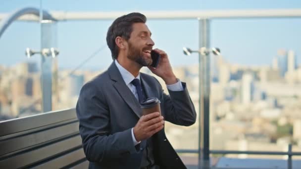 Uvolněný podnikatel mluví mobilní telefon na terase. Šťastný vousatý muž v cele s kávou na ranním panoramatu. Radostný kancelářský pracovník odpočívající na balkóně si užívá příjemného rozhovoru o přestávce - Záběry, video