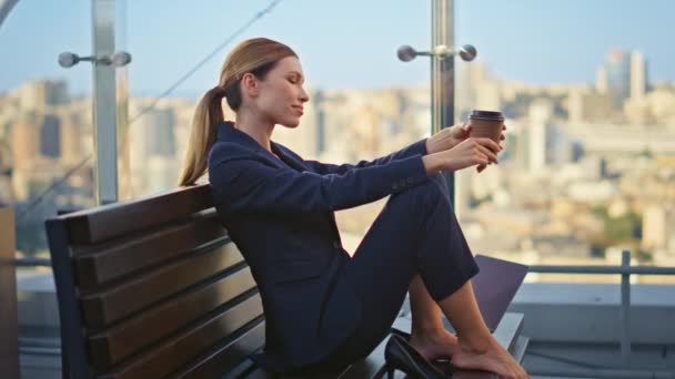 Calm manager taking break enjoying moment on glass terrace. Dreamy woman sitting bench barefoot holding coffee cup at cityscape alone. Beautiful tired businesswoman thinking resting on office balcony. - Footage, Video