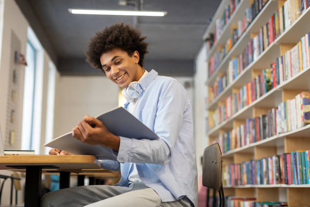 Mosolygó fekete férfi diák ül a könyvtárban, jegyzeteket ír a másolókönyvben, könyv, sugárzó vidámság a hangulatos könyvtár, szabad hely - Fotó, kép