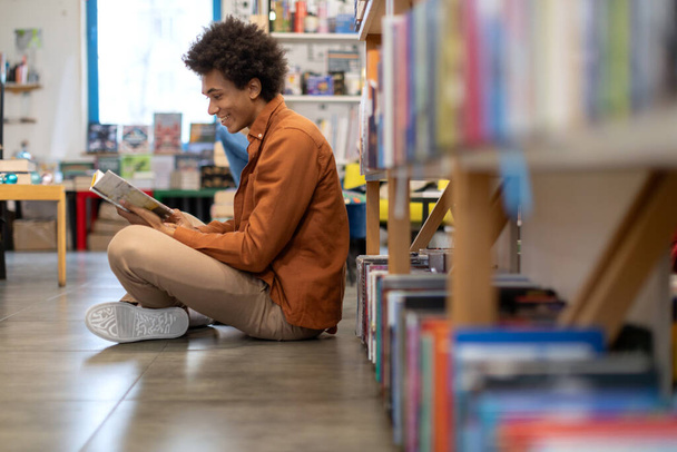 Nero studente ragazzo lettura libro mentre seduto sul pavimento vicino librerie nella biblioteca universitaria, scena serena di apprendimento immersivo e tradizionale - Foto, immagini