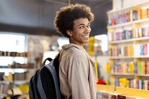Библиотечный момент запечатлен счастливым чернокожим студентом с рюкзаком, горячо улыбающимся в камеру, излучающим позитив и радость в разгар учебы - Фото, изображение