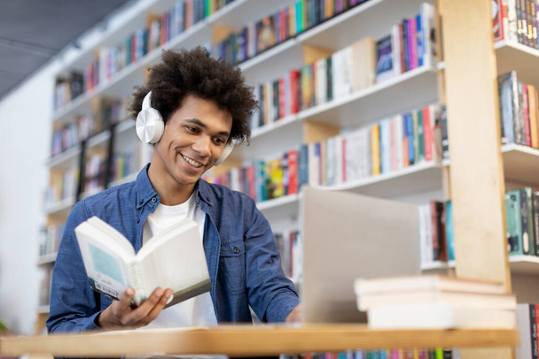 Többfunkciós fekete diák fickó ül a könyvtár asztalánál a laptop előtt, fejhallgatót visel, könyvet tart, és nézi a számítógép képernyőjét. - Fotó, kép