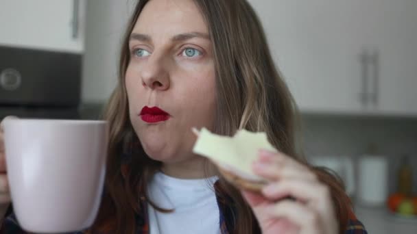 Ungesunde Kaukasierin mit roten Lippen, die in der heimischen Küche leckeres Sandwich beißt. Frauen trinken heißen Tee oder Kaffee, machen Pause. Nahaufnahme. Richtige Ernährung gesunde Fast Food ungesunde Wahl - Filmmaterial, Video