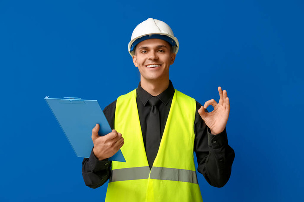 Jonge mannelijke ingenieur met klembord tonen OK op blauwe achtergrond - Foto, afbeelding