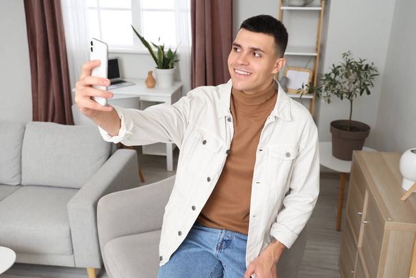 Nuori mies, jolla on matkapuhelin, ottaa selfien nojatuolilla kotona - Valokuva, kuva