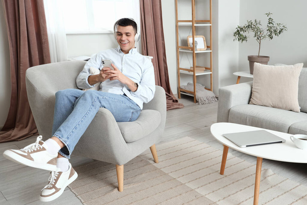 Fiatal férfi fülhallgatóval mobiltelefont használ fotelban otthon - Fotó, kép