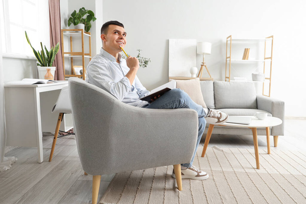 Nachdenklicher junger Mann mit Notizbuch sitzt zu Hause im Sessel - Foto, Bild