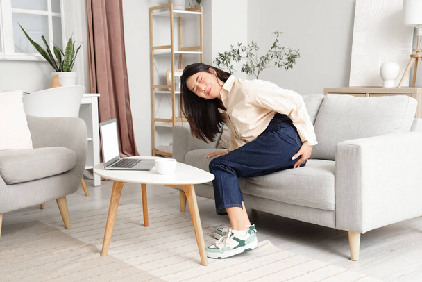 Joven mujer asiática con hemorroides sentada en el sofá en casa - Foto, imagen