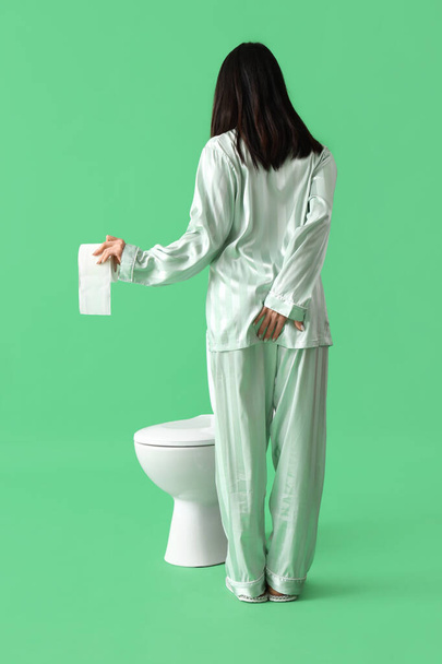 Jeune femme en pyjama avec rouleau de papier et cuvette de toilette sur fond vert, vue de dos. Concept des hémorroïdes - Photo, image