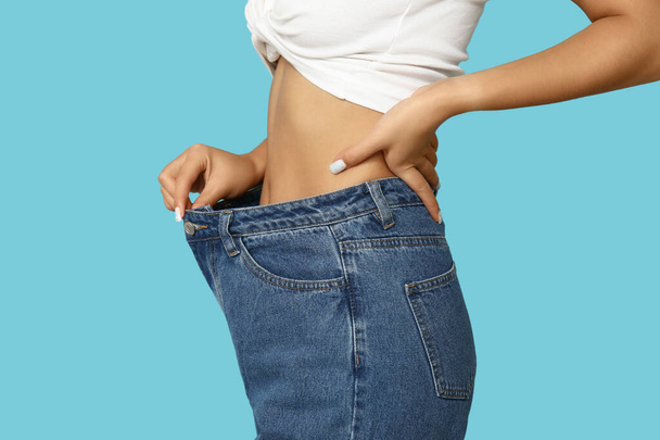 Mujer joven en pantalones vaqueros sueltos después de la pérdida de peso sobre fondo azul, primer plano - Foto, Imagen