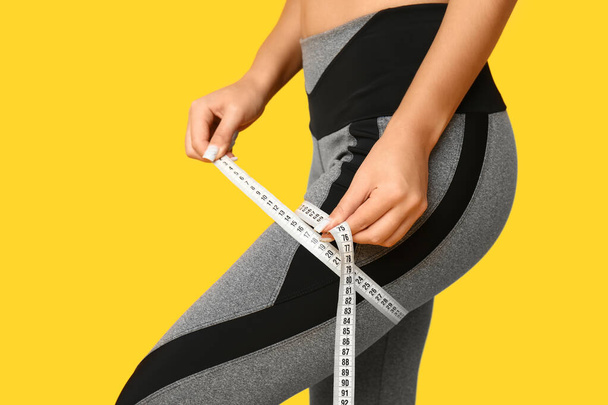 Young woman in sportswear measuring her thigh on yellow background, closeup. Weight loss concept - Photo, Image
