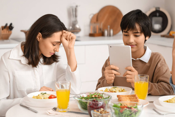 Nastolatek korzystający z tabletu i jego zdenerwowana matka przy stole podczas kolacji w kuchni. Koncepcja problemu rodzinnego - Zdjęcie, obraz