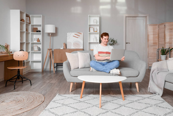 Junger Mann mit Laptop auf grauem Sofa im Wohnzimmer - Foto, Bild