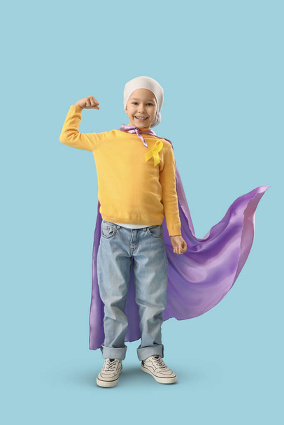 Petite fille mignonne en costume de super héros avec ruban jaune sur fond bleu. Concept de sensibilisation au cancer chez l'enfant - Photo, image