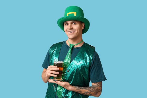 Glücklicher junger tätowierter Mann in grüner Kleidung und mit einem Glas Bier auf blauem Hintergrund. St. Patrick's Day - Foto, Bild