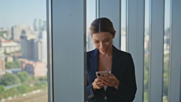 Улыбающийся работник читает сообщение на смартфоне в современном панорамном офисе. Привлекательный корпоративный менеджер в костюме наслаждается мирным перерывом просмотра мобильного телефона. Спокойная женщина-босс смотрит городской пейзаж - Кадры, видео