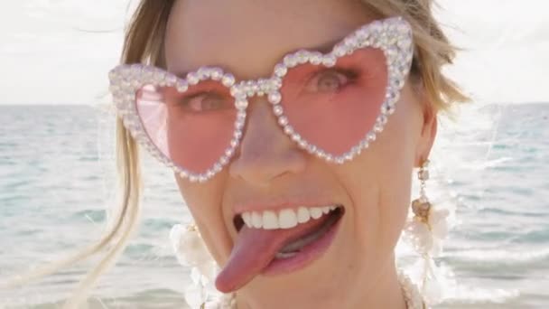 Portret van een grappige hilarische vrouw met een roze bril die domme gezichten trekt, en doet alsof ze hardop zingt op het strand. Positief meisje dat plezier heeft met tongzoenen en zoenen. Close-up emotionele bruid - Video
