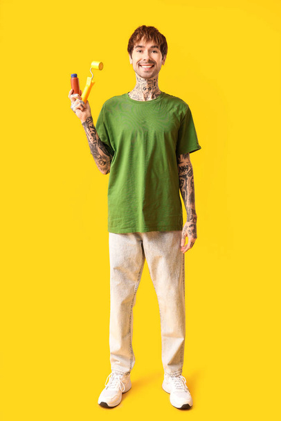 Ευτυχισμένος νεαρός άνδρας με χρώμα και ρολό σε κίτρινο φόντο - Φωτογραφία, εικόνα