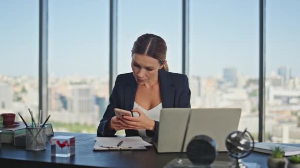 Vállalati alkalmazott ellenőrzi a mobilt az asztalnál. Elfoglalt irodai női menedzser, aki laptopot gépel üveges munkahelyen. Mosolygó üzletasszony okostelefon olvasás üzenet a közösségi média. - Felvétel, videó