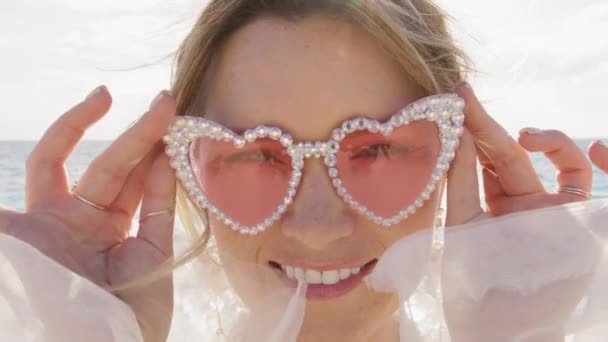 lähikuva nuori nainen vaaleanpunaisissa laseissa iskee silmää kameralle meren taustalla. Leikkisä nainen glamourin silmälaseissa katsoo kameraan. Muotokuva muoti tyttö silmää rannalla. Muoti malli vinkki silmä - Materiaali, video