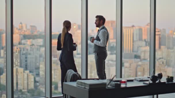 Business couple talking city view in evening. Corporate workers discussing project resting office with coffee at panoramic window. Two colleagues partners working late. Serious man with lady manager - Footage, Video