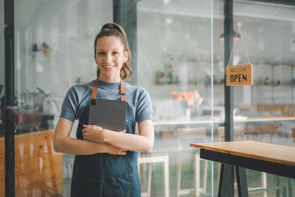 Piękna młoda baristka w fartuchu trzymająca tablet i stojąca przed drzwiami kawiarni z tabliczką z otwartym znakiem. Rozpoczęcie działalności gospodarczej przez właściciela firmy, koncepcja sprzedawcy MŚP. - Zdjęcie, obraz