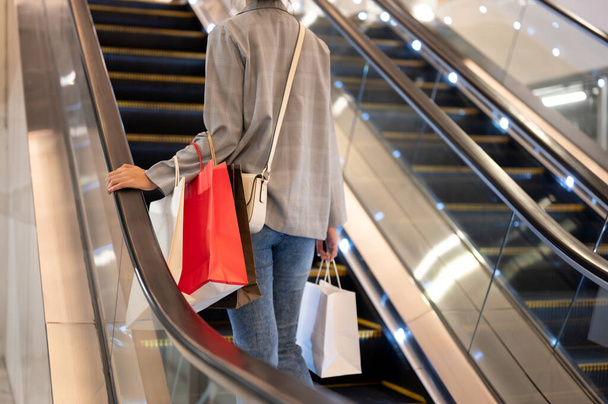 Cropped image d'une femme en vêtements à la mode avec des sacs à provisions monte un escalier roulant dans un centre commercial. vie urbaine, consumérisme, style de vie, concepts de beauté et de mode - Photo, image
