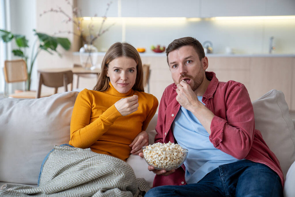 Impressed man and woman watching TV series sitting on couch at home eating popcorn. Interested family couple wife husband enjoy movie on sofa. Pastime, leisure, television show, resting, lazy holiday. - Photo, Image