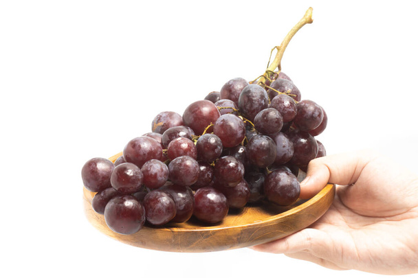Mano sosteniendo un ramo de uvas rojas orgánicas frescas deliciosa fruta en un plato de madera aislado en el camino de recorte de fondo blanco - Foto, imagen