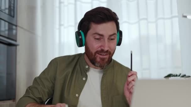 ハッピーな白人フリーランサーは,自宅のオフィスでポータブルラップトップで作業しながら楽しんでいるカジュアルな服装を着ています. ドラムティックとして鉛筆を使用してヘッドフォンの陽気な男と屋内で仮想ドラムを再生. - 映像、動画