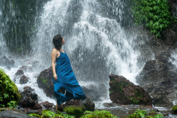 Portret van de jonge mooie vrouw die voor een waterval staat in een blauwe jurk. Bali. Indonesië. - Foto, afbeelding