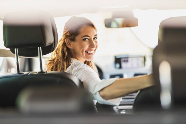 Portret van een jonge vrouw die in een auto zit en achterom over haar schouder kijkt voordat ze gaat rijden. - Foto, afbeelding