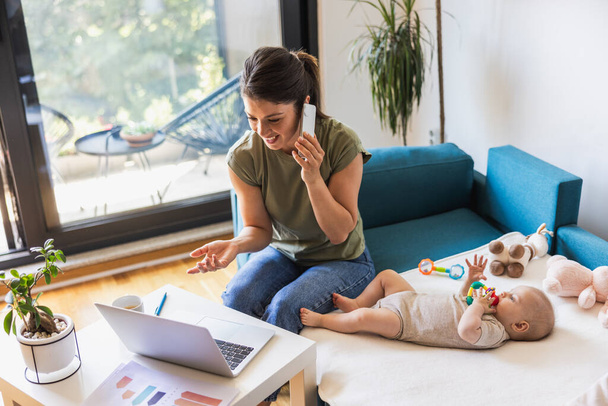 Női vállalkozó kommunikál a mobiltelefon, miközben dolgozik a laptop és vigyáz a baba otthon. - Fotó, kép