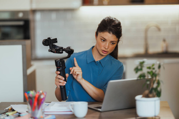 Femme vlogging sur les produits de photographie et montrant cardan pour appareil photo tout en ayant réunion en ligne sur son ordinateur portable à la maison. - Photo, image
