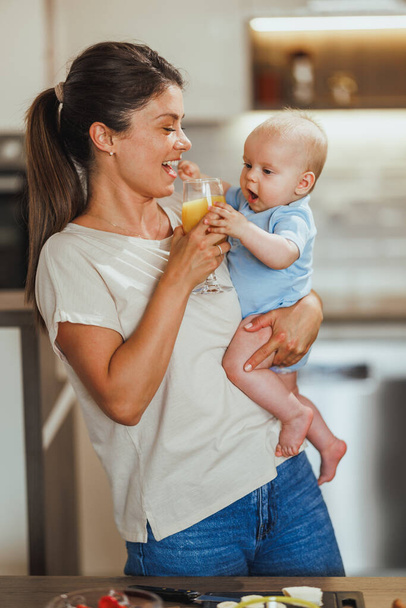Młoda troskliwa matka trzyma dziecko i szklankę soku w domu. - Zdjęcie, obraz