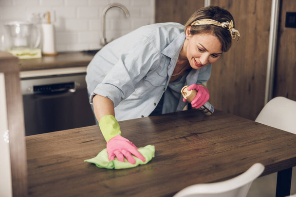 Mujer feliz manteniendo su casa limpia y limpiando una mesa de cocina en casa. Lleva guantes protectores y una botella de spray.. - Foto, Imagen