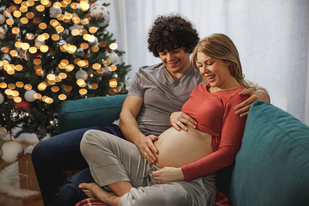 クリスマスにリビングルームでリラックスして幸せな妊婦カップル. - 写真・画像