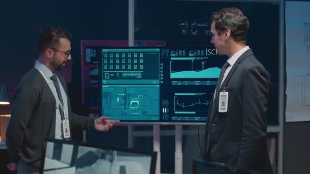 Střední záběr dvou etnicky různorodých úředníků kybernetické bezpečnosti analyzujících informace z tajného síťového programu na obrazovce, když pracují v kanceláři pozdě v noci - Záběry, video