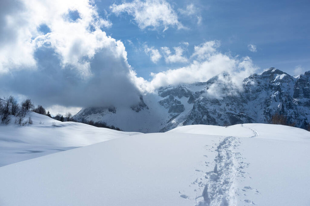 Paysage montagneux, entièrement recouvert de neige avec des traces de randonneurs sur la neige ; en arrière-plan, des montagnes enneigées au milieu des nuages - Photo, image