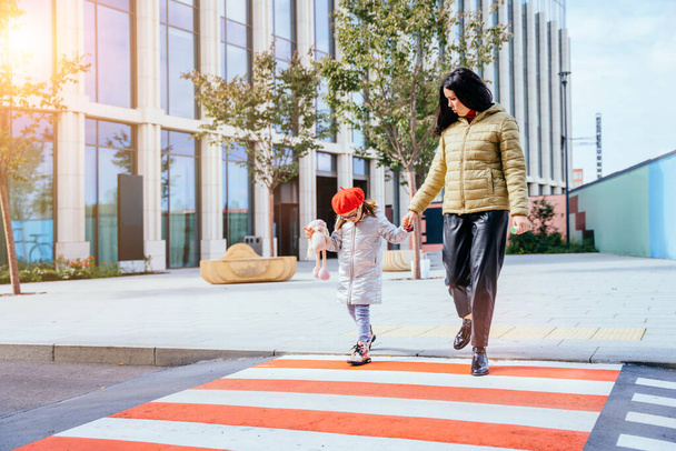 Jeune mère caucasienne traversant la rue et se rendant au centre-ville avec sa petite fille portant des lunettes et un béret rouge en automne. Pleine hauteur. - Photo, image