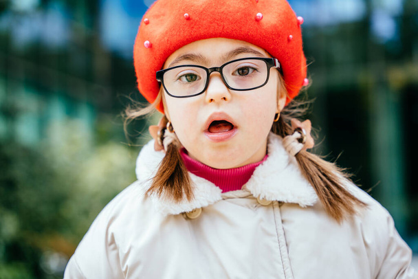 Close up outdoor portrait of cute little girl in eyeglasses with two braids wearing red beret at city street. - Photo, Image