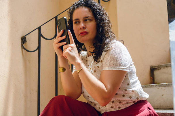 jeune femme latine avec des bigoudis, est assis sur des escaliers à l'extérieur de la maison avec téléphone portable près de son visage avec difficulté à essayer de lire l'écran, copier l'espace. - Photo, image