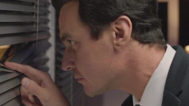 Medium closeup of serious Caucasian male FBI agent in suit looking through hole in window blind and speaking into walkie talkie while on secret mission in office at night - Footage, Video