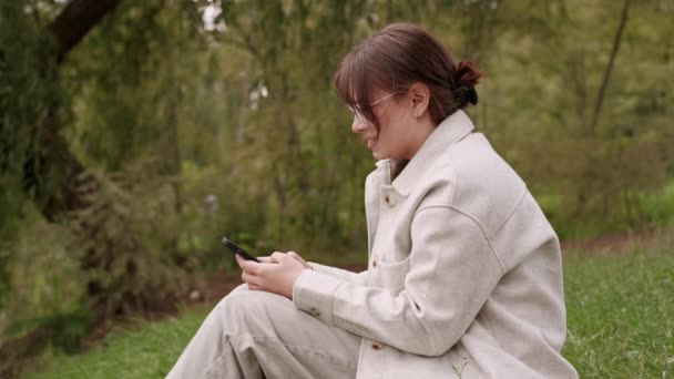 Fiatal nő ül a füvön a parkban, és gépelés okostelefonon. - Felvétel, videó