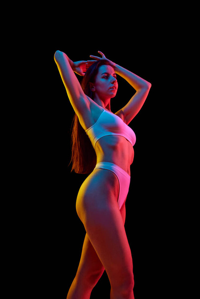 Portrait of beautiful woman with slim, fit figure in inner wear posing against black studio background in mixed neon light. Concept of wellness, weight loss, treatment, health care, fitness - Photo, Image