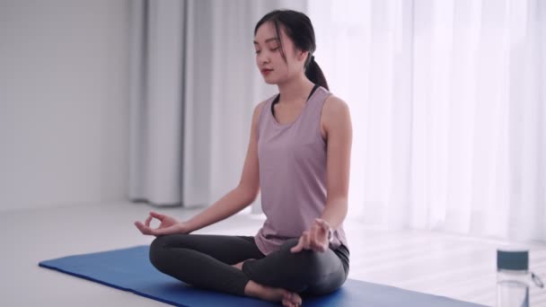 Mujer asiática en atuendo de entrenamiento practica meditación en una esterilla de yoga en su casa sala de estar. Perfecto para mostrar relajación y atención plena en un entorno doméstico. Imágenes de alta calidad 4k - Imágenes, Vídeo