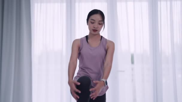 Uma mulher asiática em traje de treino graciosamente alonga seus músculos usando ioga ou poses de exercício em um tapete em suas casas sala de estar. Ideal para fitness ou promoção de ioga. - Filmagem, Vídeo