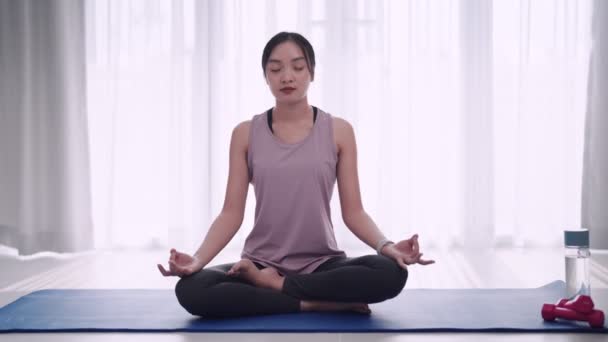 Asian woman in workout attire practices meditation on a yoga mat in her homes sitting room. Perfect for showcasing relaxation and mindfulness in a domestic setting. High quality 4k footage - Materiał filmowy, wideo