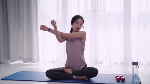 Uma mulher asiática em traje de treino graciosamente alonga seus músculos usando ioga ou poses de exercício em um tapete em suas casas sala de estar. Ideal para fitness ou promoção de ioga. Imagens 4k de alta qualidade - Filmagem, Vídeo
